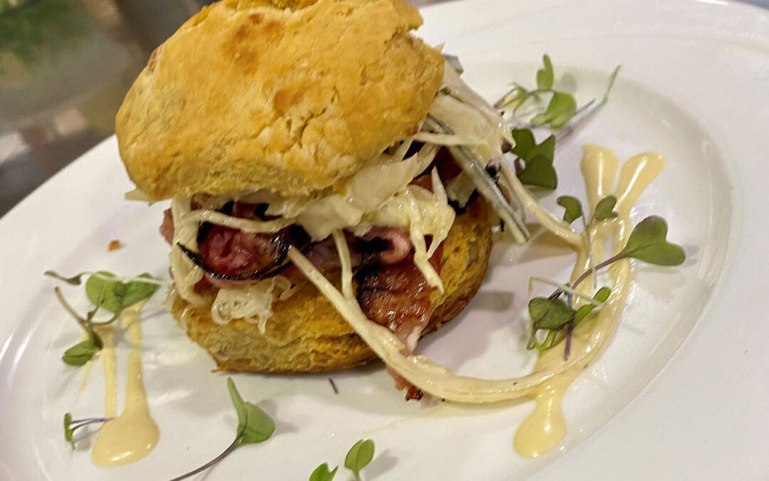 The best Pastrami Reuben Dish in Bergen County! This dish starts with Thinly Sliced Pastrami and Smoked Duck Breast and Coleslaw served on a Toasted Duck Fat Swiss Cheese Biscuit, with Hot Mustard Vinaigrette and Julienne Dill Pickle. Served on a white plate on a stainless steel table.